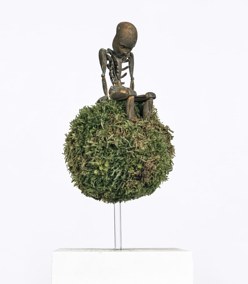 Anthony by Federico Clapis: bronze-toned skeletal child sitting with bowed head on a green moss sphere raised on a clear rod