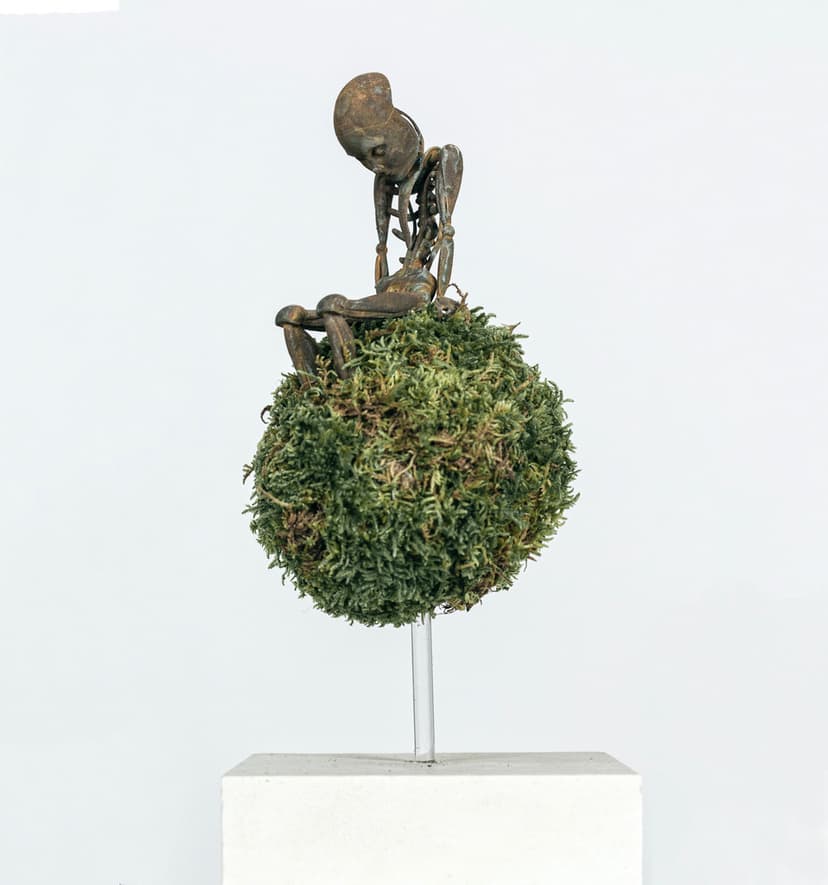 Anthony by Federico Clapis, side view: thin metal figure with a large head hunched on a moss ball above a white plinth