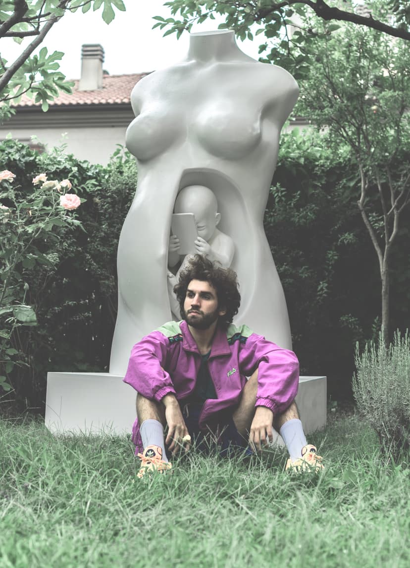 Federico Clapis in a purple jacket sits on the grass in front of his sculpture Connection, a white resin torso with a fetus inside