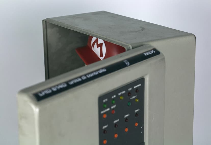 Detail of Digital Mask by Federico Clapis: half-open grey alarm control box with indicator lights, a red cap with an M visible inside
