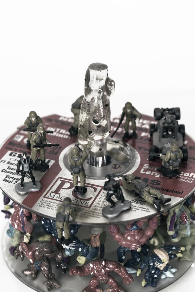 Digital war by Federico Clapis from above: miniature soldiers and a jeep on a printed PC Magazine CD around a clear resin pillar