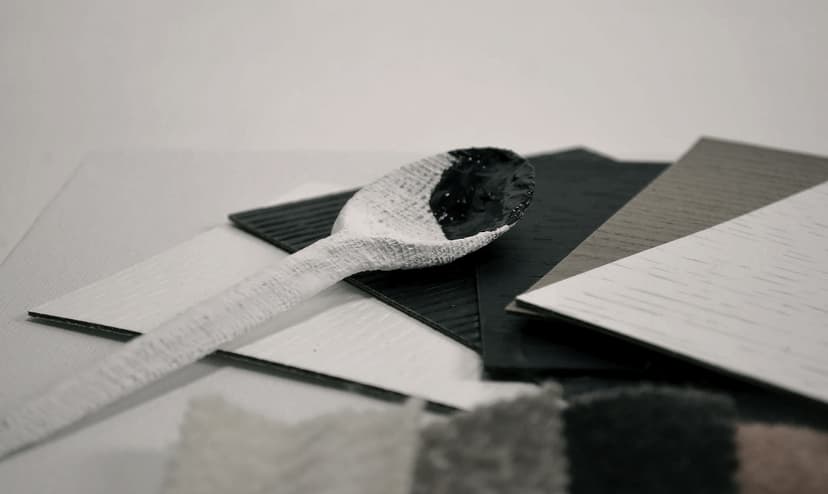 Detail of EatMateria 38 by Federico Clapis: a white textured spoon with a black-filled bowl lying on wood-grain laminate samples