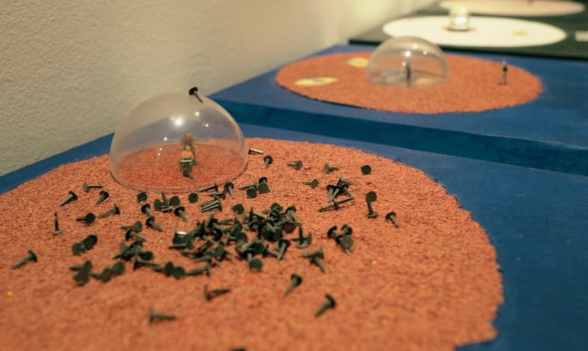 O.o.b.e by Federico Clapis: a clear dome over a tiny figure on a disc of orange grit scattered with dark nails, beside similar pieces