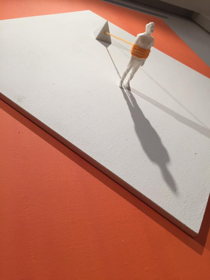 The Pyramid by Federico Clapis, angled view: white figure tied by orange wool to a pyramid on a white panel over orange canvas