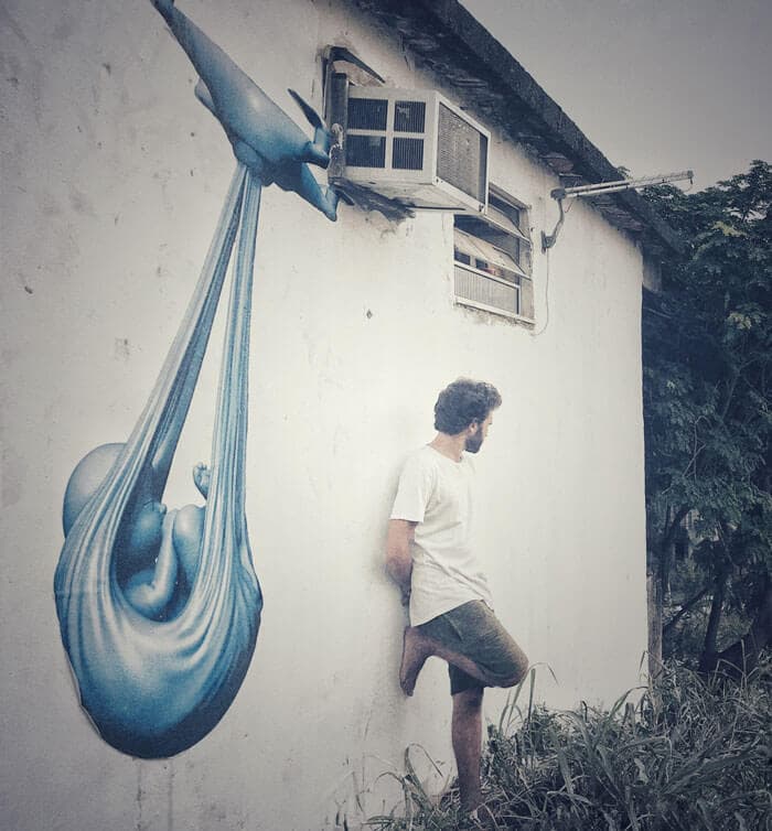Federico Clapis leans against a whitewashed wall in Cidade de Deus, Rio de Janeiro, beneath his blue Babydrone mural of a drone carrying a newborn