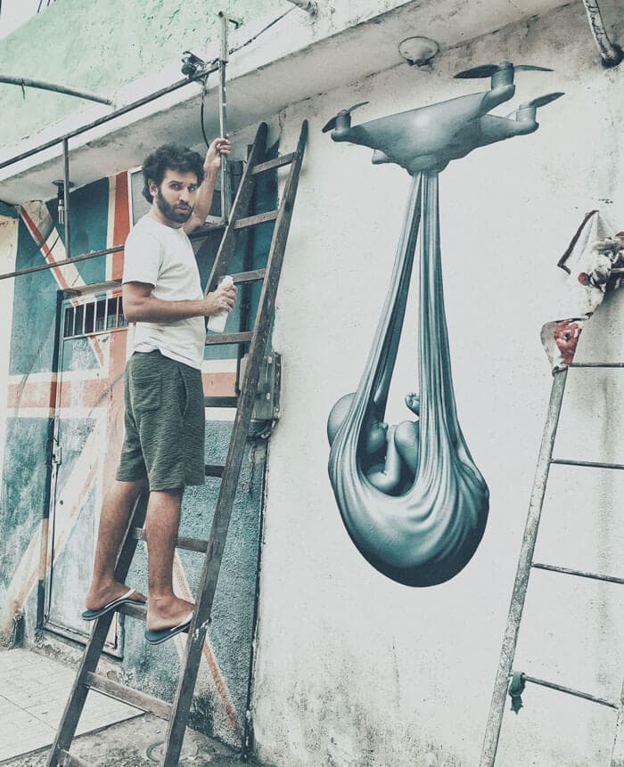 Federico Clapis on a wooden ladder with a spray can beside his grey Babydrone mural, a drone carrying a baby in a sling, City of God favela, Rio