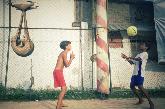 Two boys play football by a chain-link fence in Rio de Janeiro's City of God, in front of an ochre Babydrone mural by Federico Clapis