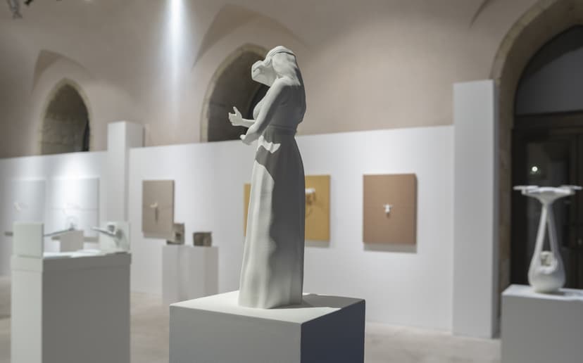 A white sculpture of a woman in a long dress stands in front of canvases with tiny figures, in Federico Clapis' Contact Zone show at EXMA