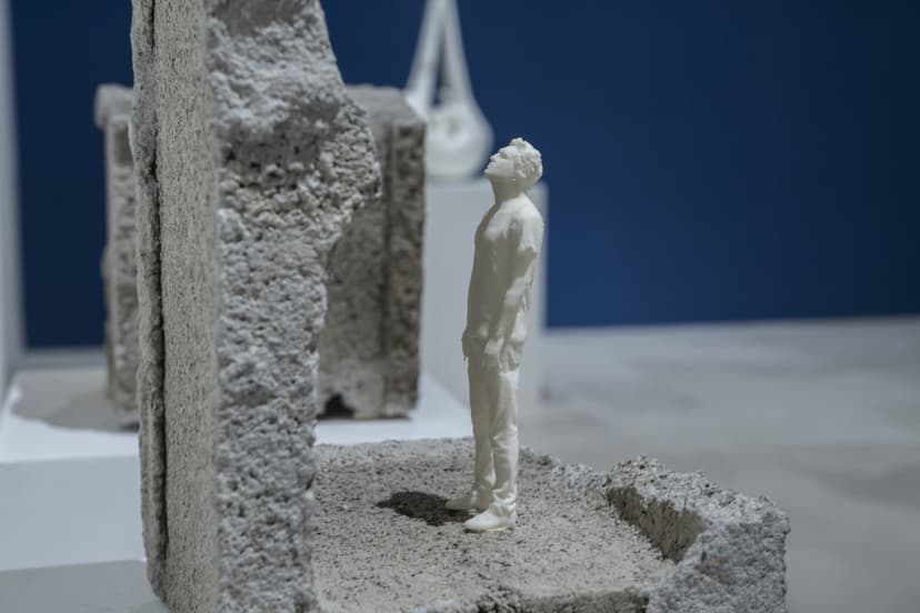 A tiny white figure looks up between broken concrete blocks, a sculpture by Federico Clapis shown against a blue wall at Contact Zone, EXMA