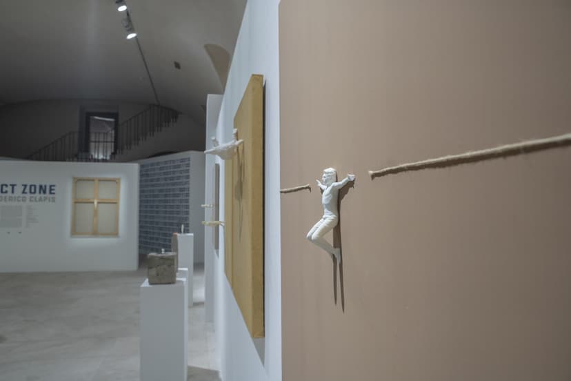 A small white figure hangs from a rope stretched across a canvas, with the Contact Zone title wall of Federico Clapis at EXMA behind