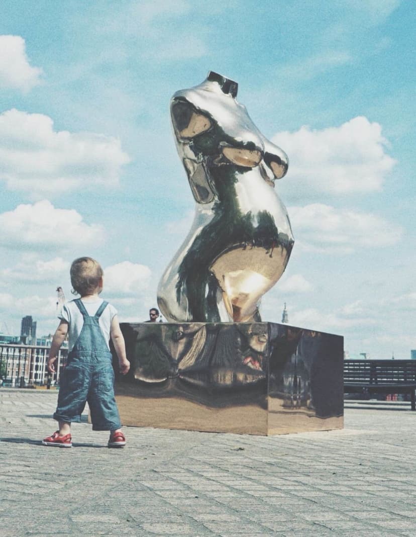 A toddler looks up at the back of Crypto Connection, Federico Clapis' polished bronze pregnant torso on a mirrored plinth, South Bank, London