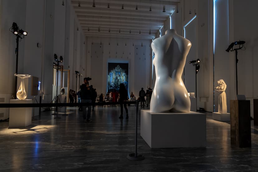 Darkened hall of Triennale Milano with white sculptures by Federico Clapis on plinths, a female torso in the foreground and visitors behind
