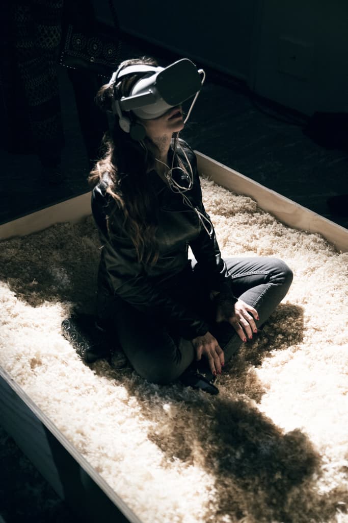 A visitor wearing a VR headset sits cross-legged in a box of wood shavings, a virtual reality installation by Federico Clapis at Triennale Milano
