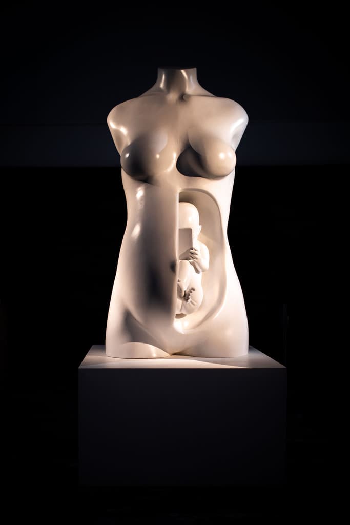 White sculpture by Federico Clapis of a female torso with a foetus holding a smartphone in the womb, lit in the dark at Triennale Milano