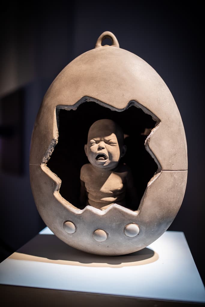 Sculpture by Federico Clapis of a crying baby inside a cracked egg-shaped shell, displayed on a white plinth at the Deep Scrolling Experience