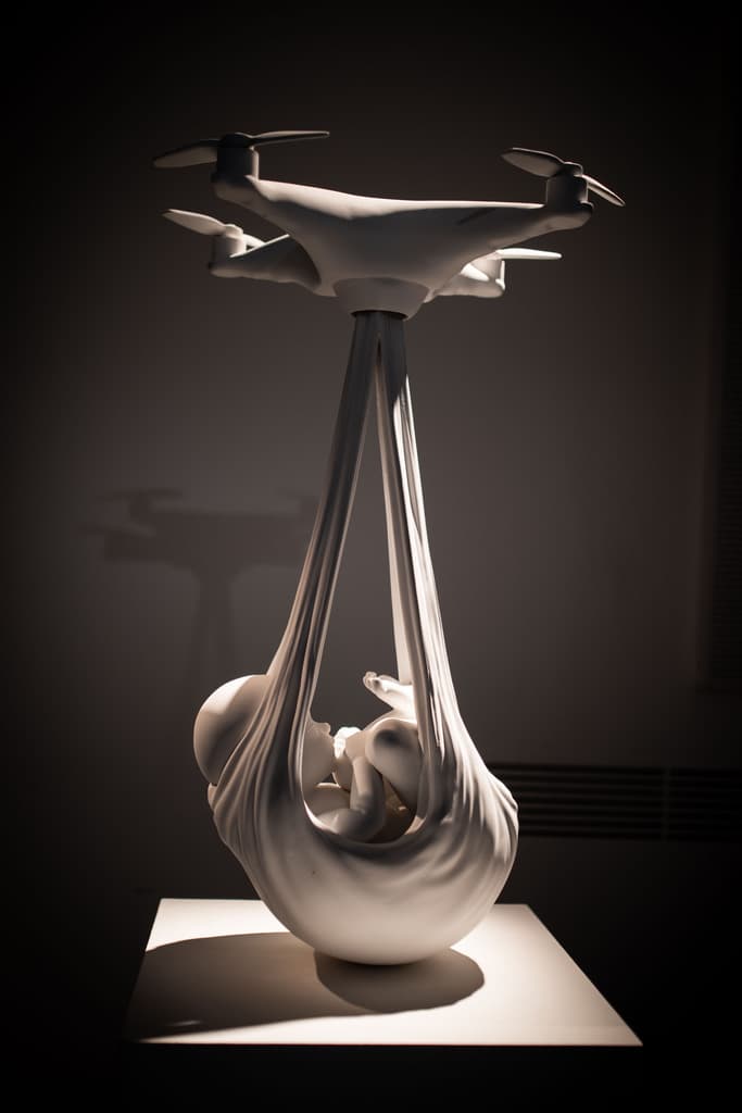 White sculpture by Federico Clapis of a newborn carried in a cloth sling by a drone, shown in the dark at Triennale Milano