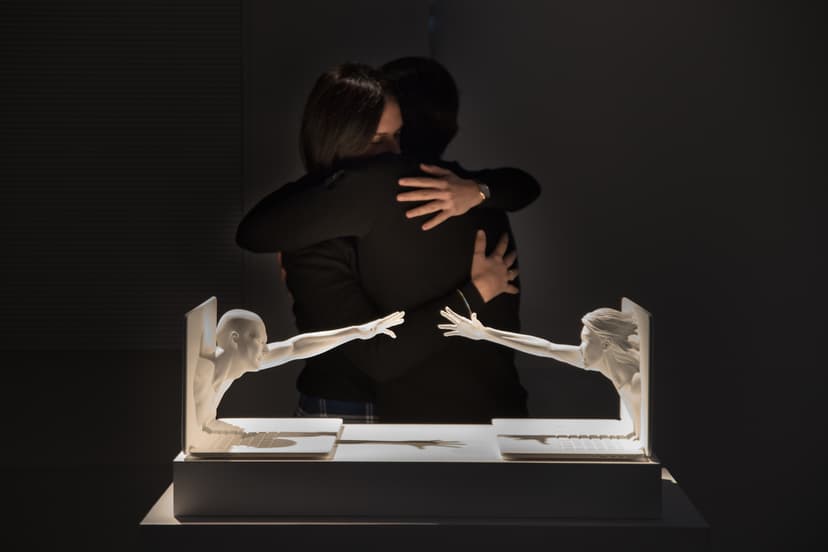 Two people embrace behind a sculpture by Federico Clapis of two figures reaching out of laptop screens, at Triennale Milano