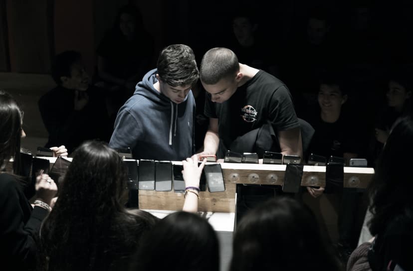 Young visitors gather around a wooden bar lined with smartphones, an installation by Federico Clapis at Triennale Milano