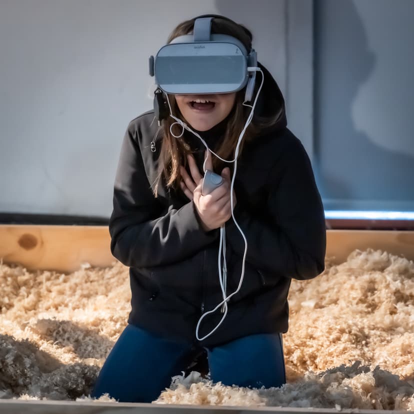 A girl in a VR headset kneels in wood shavings and reacts with surprise, inside a virtual reality installation by Federico Clapis at Triennale Milano
