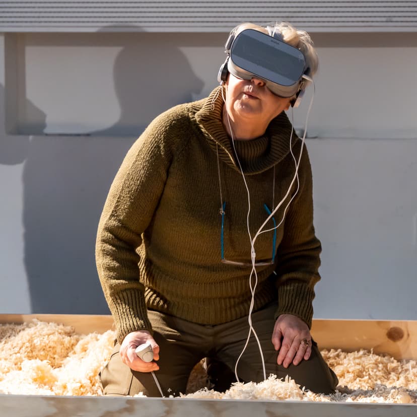 An older woman in a VR headset kneels in a box of wood shavings, experiencing an installation by Federico Clapis at the Deep Scrolling Experience