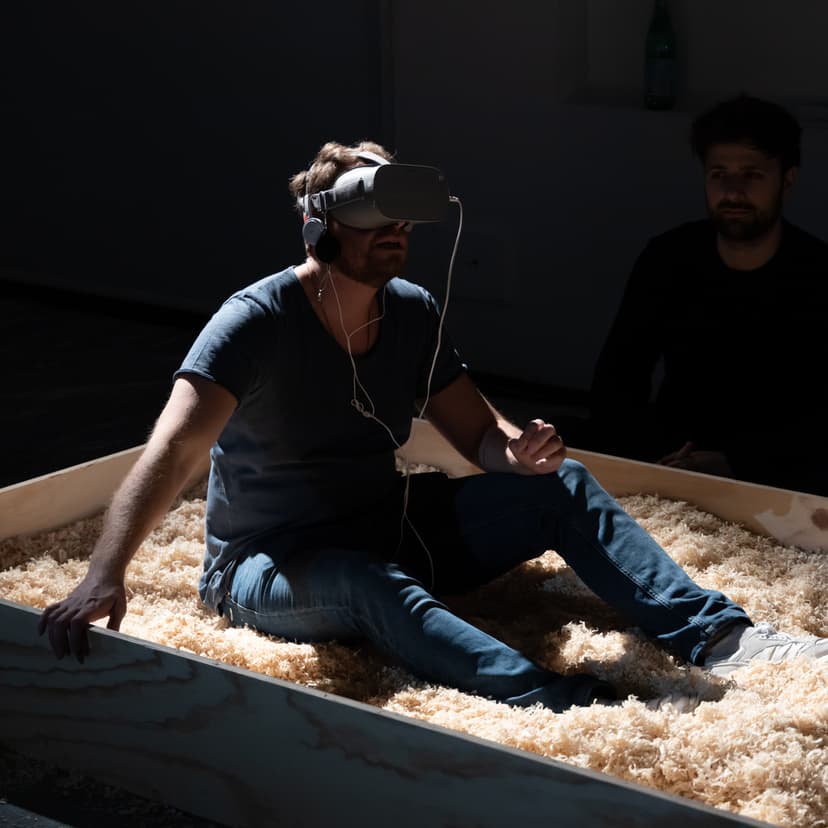 A man wearing a VR headset sits in wood shavings inside a wooden enclosure, a virtual reality work by Federico Clapis at Triennale Milano