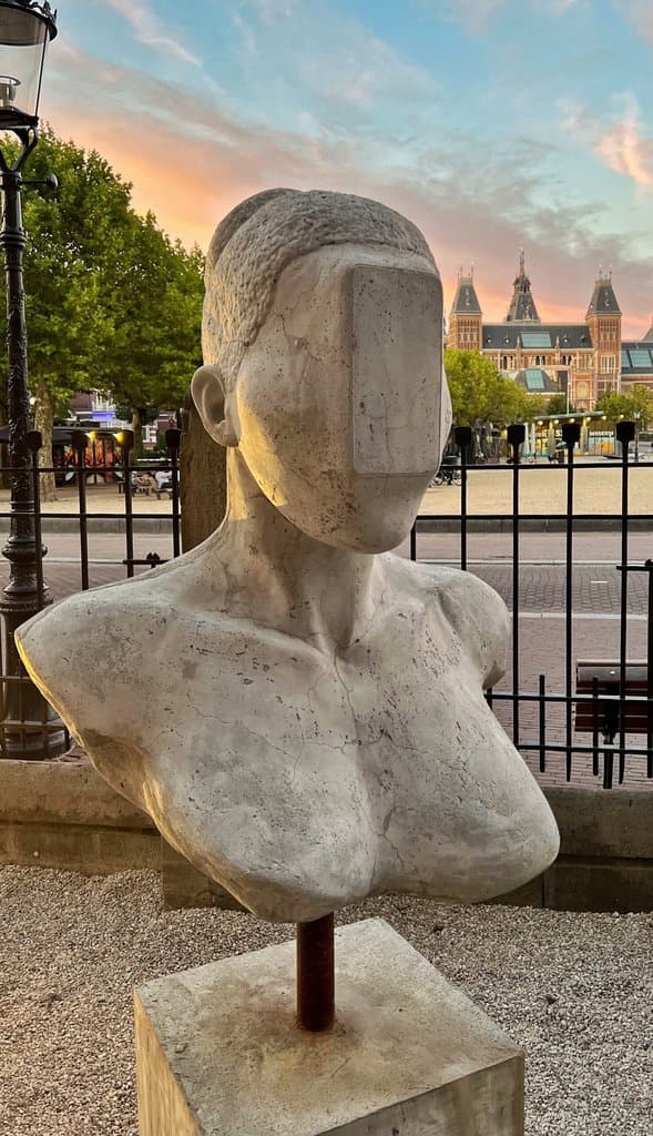 Close-up of a New Race bust by Federico Clapis, its face hidden by a smooth slab, in the Moco Museum Amsterdam garden at sunset