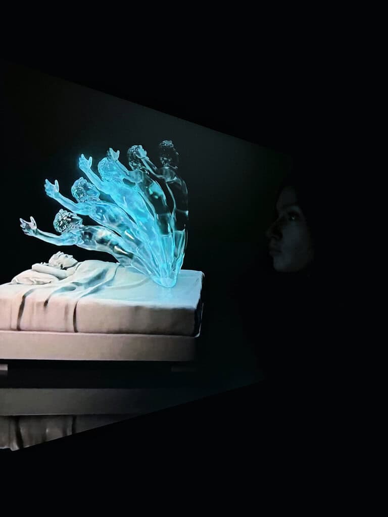Screen showing a work by Federico Clapis: glowing translucent figures rise from a sleeper on a bed, watched by a visitor at The Future of Art, NFT Paris