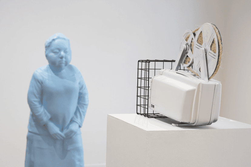 Pale blue figure of a woman with clasped hands near a white film projector with a wire basket, works by Federico Clapis at Bianchi Zardin