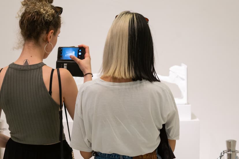 Two visitors photograph Federico Clapis' white sculpture with a phone in a bright gallery of Triennale Milano during the Festival dell'Amore